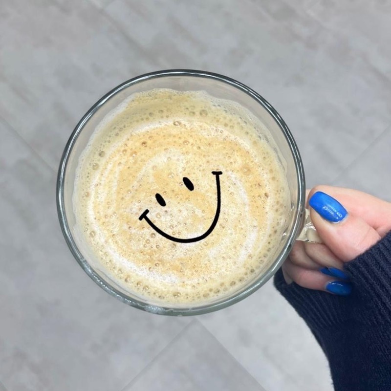 A glass mug of frothy coffee with a smiley face drawn on the foam, held in a hand with blue nails.